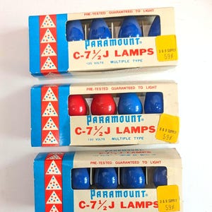 May include: Three rectangular boxes of Paramount C-7 1/2 J lamps. Each box features a blue side panel with red triangles containing blue dots. The boxes contain blue or red glass bulbs, labeled 120 volts, multiple type, and pre-tested.