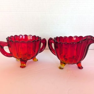 May include: A pair of ruby red glass vessels, consisting of a sugar bowl with two handles and a creamer with a single pouring spout. Both pieces feature a scalloped rim, vertical ribbed detailing, and three small feet with amber-toned bases.