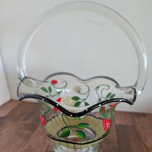 May include: A clear glass basket with a handle, featuring hand-painted strawberries and green leaves. The rim is scalloped and outlined with black and white polka dots. The basket sits on a pedestal base.