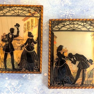 May include: Two framed silhouette art pieces. Each features a black silhouette of a couple in a romantic scene, set against a backdrop of a European town. The frames have a decorative border with a geometric design.