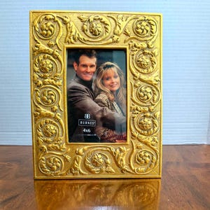 May include: Ornate gold-colored picture frame with a rectangular shape. The frame features detailed floral and scrollwork designs. The frame holds a 4x6 inch photo of a couple. The brand name "Burnes" is visible on the frame.