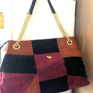 May include: A patchwork suede tote bag with a gold chain strap. The bag features a geometric design in shades of brown, black, and burgundy. A small gold emblem of a dog is on the front.
