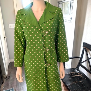 May include: A green coat featuring a white polka dot pattern, a wide collar, long sleeves, and three front toggle closures with metallic hardware. The coat has a straight silhouette and reaches mid-thigh length on the mannequin.