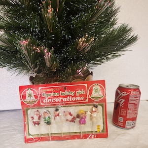 May include: A package of Commodore figurine holiday pick decorations with various Christmas characters. A small, artificial Christmas tree and a red Coca-Cola can are also in the image.