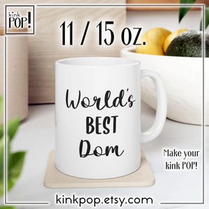 Puede incluir: Taza de cerámica blanca con texto negro que dice "World's Best Dom" sobre un posavasos blanco. La taza es de 11/15 oz.
