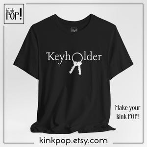 Puede incluir: Camiseta negra con la palabra "Keyholder" en blanco, con un llavero y dos llaves. La camiseta también tiene el texto "Make your kink POP!" y la URL de la tienda Etsy.