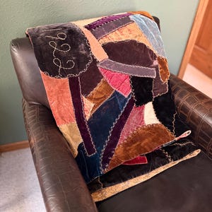 May include: A patchwork quilt made of various velvet fabric scraps in shades of purple, blue, red, brown, and tan. The quilt features visible white decorative stitching along the seams and the embroidered text 1922 on a dark purple velvet patch.