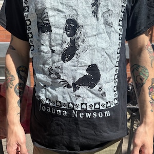 Może przedstawiać: Czarny T-shirt z białym nadrukiem graficznym przedstawiającym twarz kobiety i tekstem "Joanna Newsom".