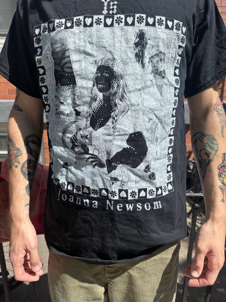 Może przedstawiać: Czarny T-shirt z białym nadrukiem graficznym przedstawiającym twarz kobiety i tekstem "Joanna Newsom".