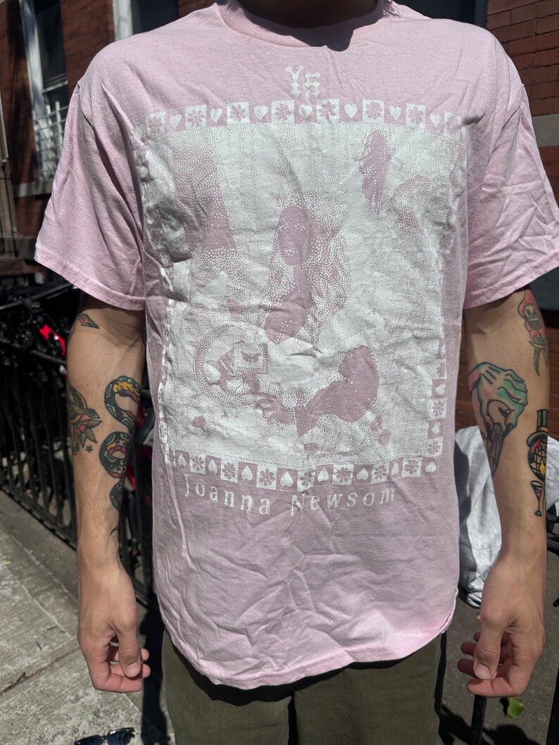 Może przedstawiać: R&oacute;żowy t-shirt z białym nadrukiem graficznym przedstawiającym kobietę z koroną i tekstem "YS" i "Joanna Newsom".