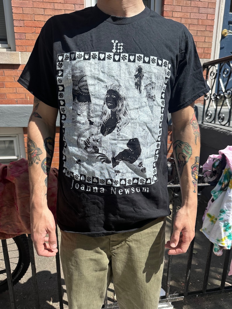Może przedstawiać: Czarny T-shirt z białym nadrukiem graficznym przedstawiającym kobietę z długimi włosami i tekstem "YS" i "Joanna Newsom".