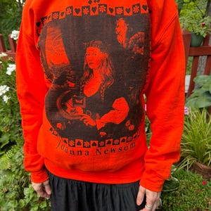 May include: A bright orange crewneck sweatshirt featuring a dark brown graphic print on the front. The design includes the text YS at the top, Joanna Newsom at the bottom, and a central illustration framed by a border of hearts and flowers.