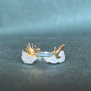 Könnte beinhalten: Ein silberner Ring mit einem goldenen Sonnen- und Vogeldessin. Die Sonne hat eine strukturierte Oberfläche und befindet sich in der Mitte des Rings. Der Vogel befindet sich auf der rechten Seite, und der Ring hat ein offenes Design.