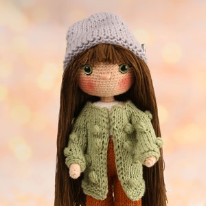 May include: Handmade doll with long brown hair, wearing a gray knit hat, a green knit jacket with textured details, and orange pants. The doll has a crocheted face with rosy cheeks and green eyes. A charming, handcrafted item.