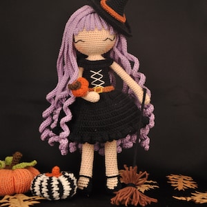 May include: A crocheted witch doll with purple hair, a black dress, and a black witch hat with an orange band. The doll is holding a small orange pumpkin and a broom. The doll is standing on a black surface with two pumpkins and some fall leaves.