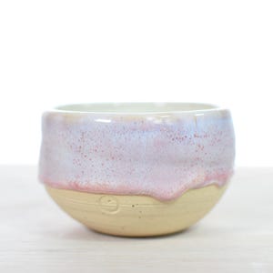 Pode incluir: Uma pequena tigela de cerâmica com base arredondada e abertura larga. A parte inferior da tigela apresenta um acabamento em argila bege natural, enquanto a parte superior é revestida com um esmalte brilhante e escorrido em rosa pastel, lavanda e branco.