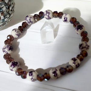 May include: A beaded bracelet featuring alternating round white ceramic beads with purple floral patterns and smaller, faceted translucent dark brown glass beads arranged in a circular pattern on a white surface.