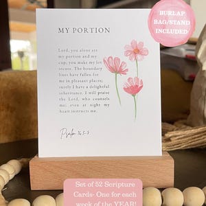 May include: A white rectangular card featuring the text MY PORTION and Psalm 16:5-7 with a watercolor illustration of three pink flowers. The card is displayed in a light-colored wooden block stand. A pink circular graphic states BURLAP BAG/STAND INCLUDED.