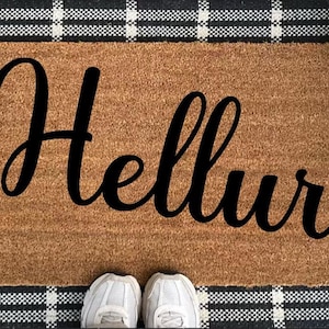 Könnte beinhalten: Eine rechteckige Fußmatte mit dem Wort "Hellur" in großer, schwarzer Schreibschrift. Die Matte ist braun mit einem schwarz-weiß karierten Rand. Weiße Turnschuhe sind unten zu sehen.