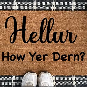Könnte beinhalten: Eine rechteckige Fußmatte aus braunem Kokosfasermaterial mit dem Text "Hellur How Yer Dern?" in schwarzer Schrift. Die Matte hat einen schwarz-weiß karierten Rand. Weiße Turnschuhe sind unten.