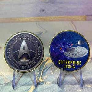 May include: Two round collectible coins on clear stands. One coin features the Star Trek Enterprise logo and the text "ENTERPRISE COLLECTION". The other coin shows the Enterprise ship in space with the text "ENTERPRISE 1701-G".