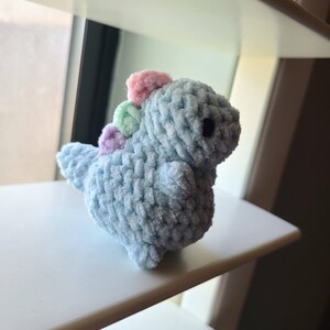 以下が含まれることがあります： ふわふわとした質感の、小さな水色の編みぐるみ恐竜のおもちゃ。恐竜はピンク、緑、紫のカラフルな背びれが並んでいます。おもちゃは白い棚の上に置かれています。