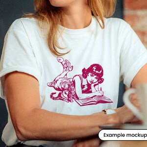 Puede incluir: Una camiseta blanca con una ilustraci&oacute;n rosa de una mujer tumbada con un gato y un libro. La mujer lleva un vestido y tiene el pelo recogido. El gato est&aacute; enroscado a su lado. El libro est&aacute; abierto y la mujer sostiene un bol&iacute;grafo.