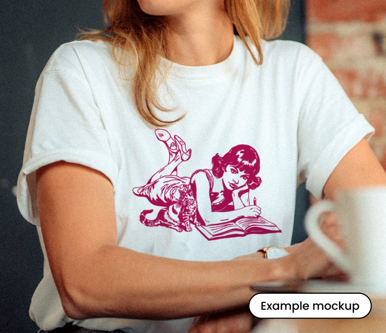 Puede incluir: Una camiseta blanca con una ilustraci&oacute;n rosa de una mujer tumbada con un gato y un libro. La mujer lleva un vestido y tiene el pelo recogido. El gato est&aacute; enroscado a su lado. El libro est&aacute; abierto y la mujer sostiene un bol&iacute;grafo.