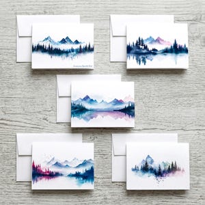 May include: Set of five watercolor landscape greeting cards with matching white envelopes. Each card features a different mountain and forest scene in shades of blue, purple, and teal. The cards are perfect for sending notes or as art.