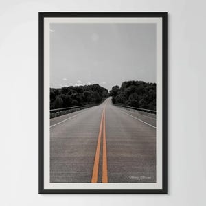 May include: A black and white photograph of a long, straight road with two orange lines down the center. The road leads to a forest in the distance.