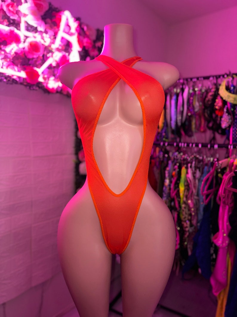 Op de afbeelding: Een feloranje bodysuit met een hoog uitgesneden beenontwerp, een diepe centrale uitsnijding en een halterhalslijn met gekruiste bandjes over de borst. Het kledingstuk is gemaakt van een doorschijnende, lichtgewicht stof met afgewerkte randen.