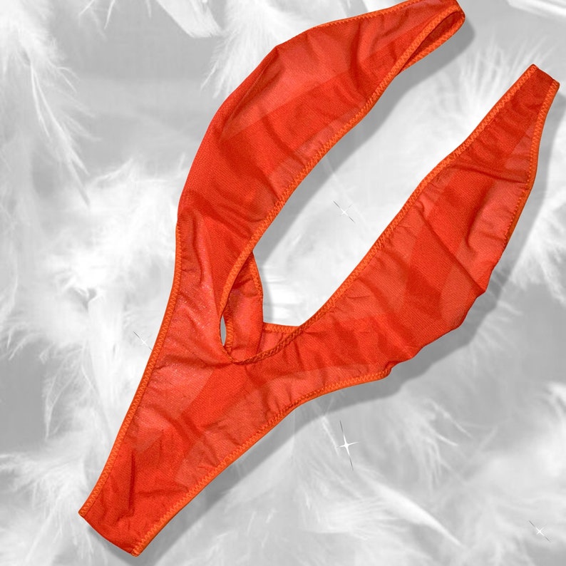 Op de afbeelding: Een feloranje string gemaakt van een doorschijnende, lichtgewicht stof. Het kledingstuk heeft een hoog uitgesneden ontwerp met dunne, flexibele zijbanden en een centrale opening, met een semi-transparante textuur en afgewerkt met bijpassende oranje stiksels.