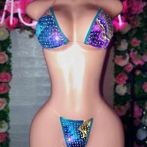 May include: A two-piece bikini set with a blue, purple, and gold design. The top is a triangle style with clear straps, and the bottom is a thong style. The bikini is embellished with rhinestones.
