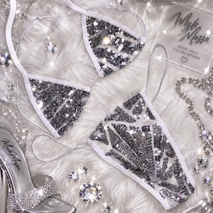 May include: A two-piece bikini set featuring white fabric adorned with silver sequins in a geometric pattern. The top has triangle cups with thin white straps, and the bottom is a matching piece. A clear high-heeled shoe with rhinestones is visible.