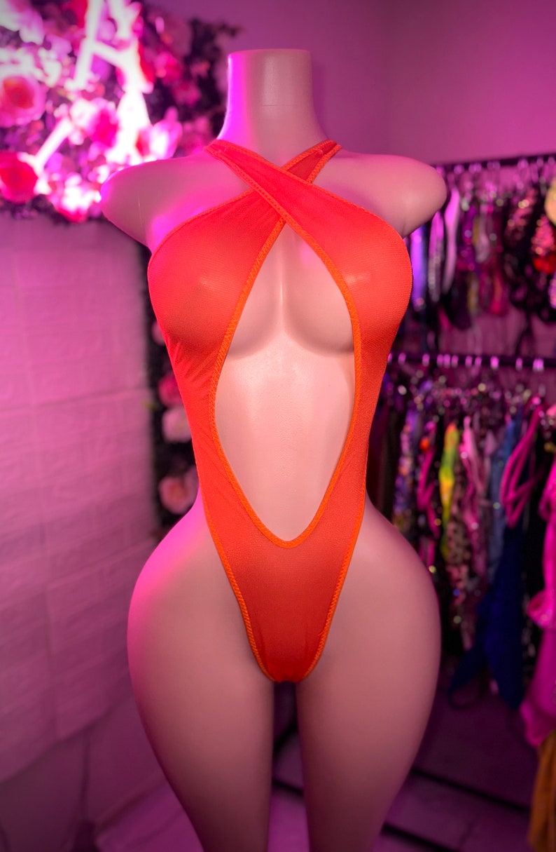 Op de afbeelding: Een feloranje bodysuit met een halterhalslijn met gekruiste bandjes op de borst, een diepe uitsnijding die doorloopt tot aan de taille en een hoog uitgesneden beenontwerp. Het kledingstuk is gemaakt van een doorschijnende, fijne mesh-stof.