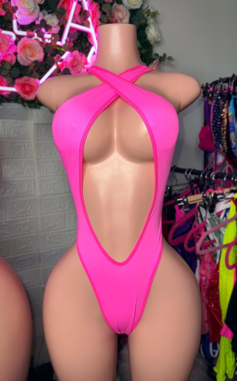 Puede incluir: Un body de una pieza en color rosa ne&oacute;n con dise&ntilde;o de pierna alta, un corte profundo que se extiende desde el escote hasta la cintura y un cuello tipo halter con tiras cruzadas que se aseguran alrededor del cuello y la parte superior del pecho.