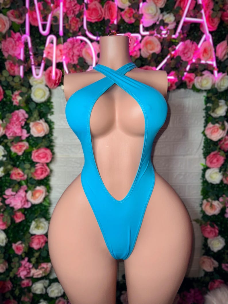 Puede incluir: Un traje de ba&ntilde;o de una pieza en color turquesa brillante con un escote profundo y un dise&ntilde;o de tirantes cruzados estilo halter en el pecho. La prenda presenta una abertura de pierna alta y una confecci&oacute;n en tela lisa de color s&oacute;lido.