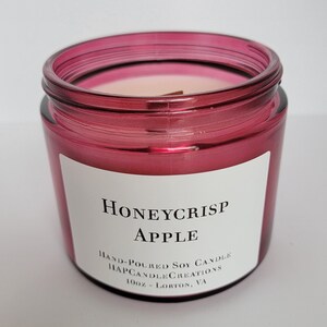 May include: A red glass jar candle with a white label that reads "Honeycrisp Apple" and "Hand-Poured Soy Candle Happy Candle Creations 10oz - Lorton, VA".