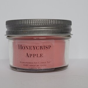 May include: A small glass jar candle with a silver lid. The candle is pink and has the text "Honeycrisp Apple" and "Hand-poured Soy Candle 5oz" on the label.