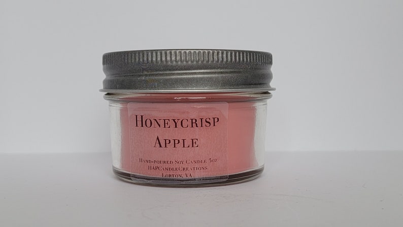 May include: A small glass jar candle with a silver lid. The candle is pink and has the text "Honeycrisp Apple" and "Hand-poured Soy Candle 5oz" on the label.