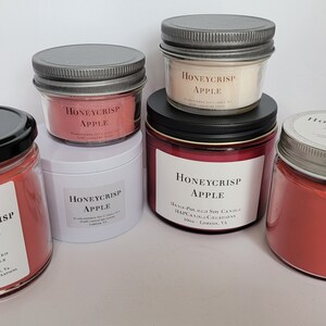 May include: Four glass jars with red soy candles. The jars have silver lids and white labels with the text "Honeycrisp Apple" printed on them. The candles are hand-poured soy candles.