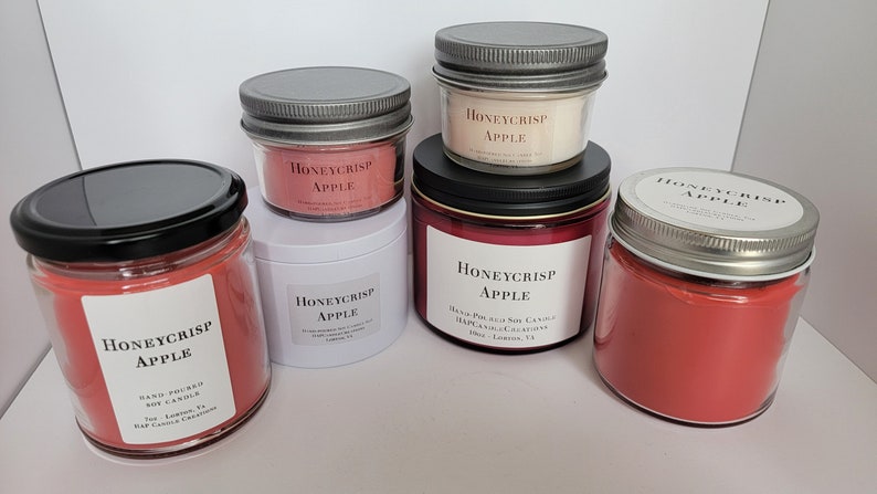 May include: Four glass jars with red soy candles. The jars have silver lids and white labels with the text "Honeycrisp Apple" printed on them. The candles are hand-poured soy candles.