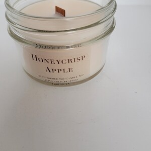 May include: A clear glass jar candle with a beige wax and a wooden wick. The label reads "Honeycrisp Apple" and "Hand-poured Soy Candle 7 oz. Mapcandlecreations Lorton, VA."
