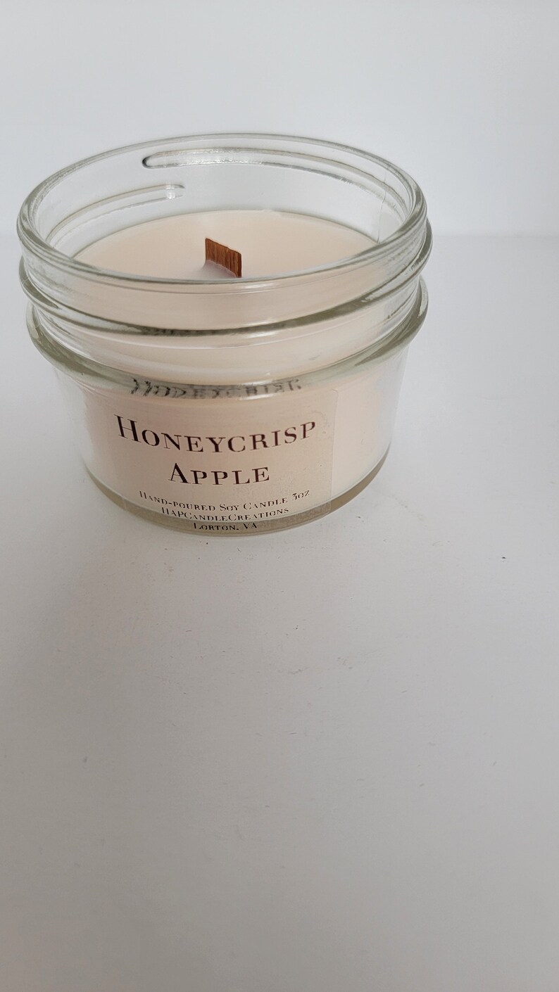 May include: A clear glass jar candle with a beige wax and a wooden wick. The label reads "Honeycrisp Apple" and "Hand-poured Soy Candle 7 oz. Mapcandlecreations Lorton, VA."
