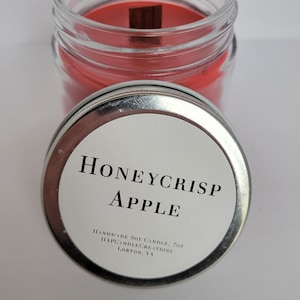 May include: A silver lid with a white label that reads "Honeycrisp Apple" and "Handmade Soy Candle, 7oz. HappyCandleCreations, Lorton, VA". The lid is on top of a glass jar with a red soy candle inside.