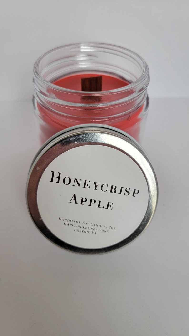 May include: A silver lid with a white label that reads "Honeycrisp Apple" and "Handmade Soy Candle, 7oz. HappyCandleCreations, Lorton, VA". The lid is on top of a glass jar with a red soy candle inside.