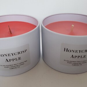 May include: Two white candle jars with red wax and a single wick. The jars are labeled "Honeycrisp Apple" and have text that reads "Hand-poured Soy Candle Jar, Happy Candle Creations, Loxton, VA".