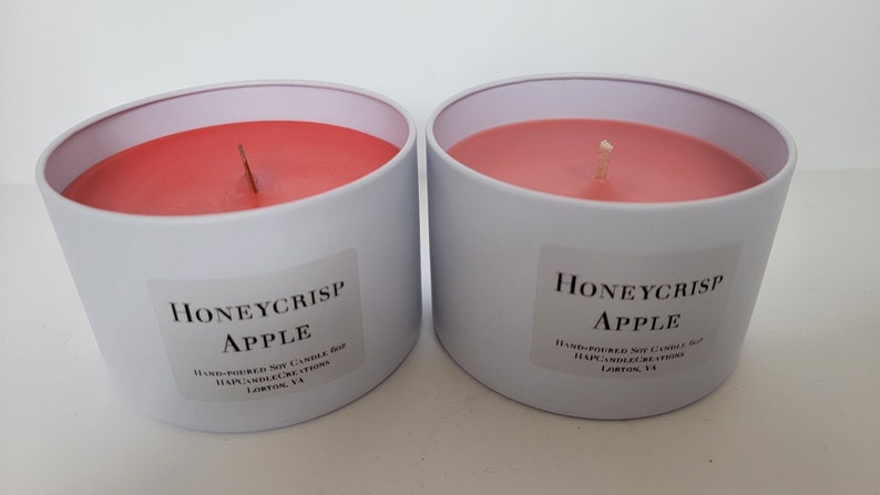 May include: Two white candle jars with red wax and a single wick. The jars are labeled "Honeycrisp Apple" and have text that reads "Hand-poured Soy Candle Jar, Happy Candle Creations, Loxton, VA".