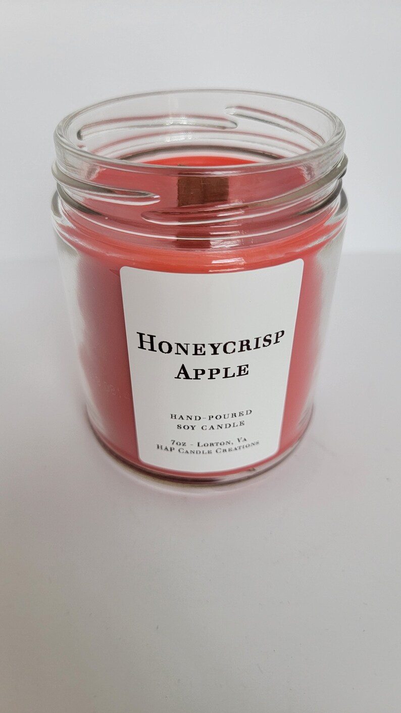 May include: A clear glass jar candle with a red soy wax scent labeled "Honeycrisp Apple". The label also states "Hand-poured Soy Candle" and "7oz - Lorton, VA. Hap Candle Creations."