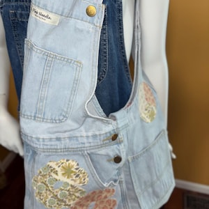 May include: Light blue denim overalls with floral patches. The overalls have adjustable straps and a large pocket on the front. The brand label reads "Pink Needle, Made in China."